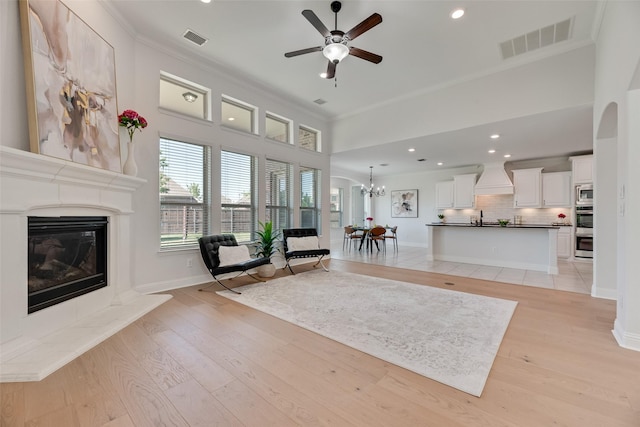 living area featuring visible vents, ornamental molding, a glass covered fireplace, and ceiling fan with notable chandelier