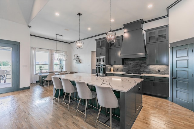 kitchen with custom exhaust hood, backsplash, light stone counters, an island with sink, and appliances with stainless steel finishes