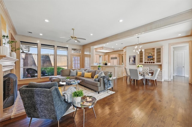 living area with recessed lighting, ceiling fan with notable chandelier, wood finished floors, crown molding, and a fireplace