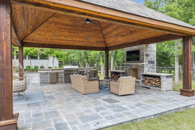 view of patio featuring an outdoor kitchen, an outdoor stone fireplace, a ceiling fan, area for grilling, and a gazebo