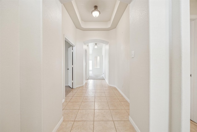 corridor featuring arched walkways, a tray ceiling, baseboards, and light tile patterned floors