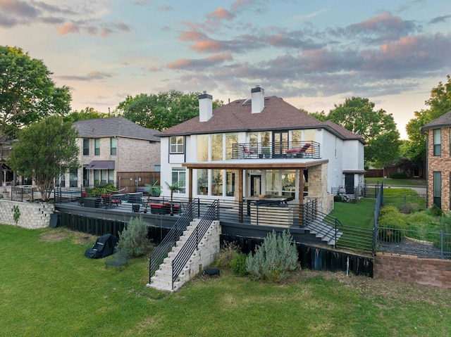 back of house at dusk featuring a chimney, fence, a lawn, stairs, and a balcony