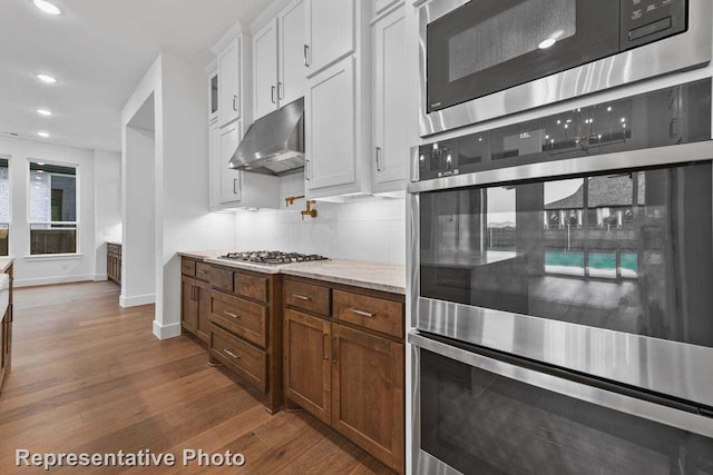 kitchen featuring dark wood finished floors, baseboards, white cabinetry, under cabinet range hood, and appliances with stainless steel finishes