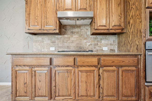 kitchen featuring brown cabinetry, decorative backsplash, gas cooktop, light countertops, and under cabinet range hood