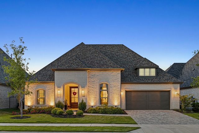 french country inspired facade featuring decorative driveway, a front lawn, an attached garage, brick siding, and roof with shingles