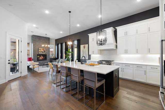 kitchen with light countertops, premium range hood, a kitchen island with sink, a fireplace, and dark wood-style flooring