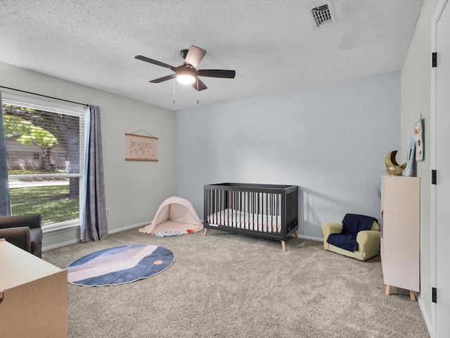 carpeted bedroom with a textured ceiling, visible vents, baseboards, a crib, and a ceiling fan