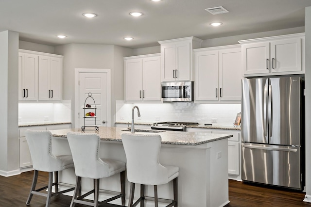 kitchen featuring dark wood-style floors, stainless steel appliances, backsplash, a center island with sink, and white cabinetry