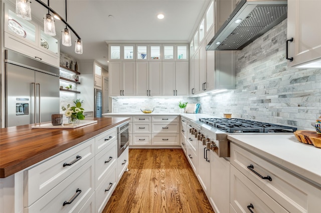kitchen with ventilation hood, light wood-style flooring, tasteful backsplash, built in appliances, and wooden counters