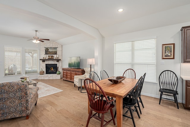 dining space with ceiling fan, vaulted ceiling, a stone fireplace, and light wood-style flooring