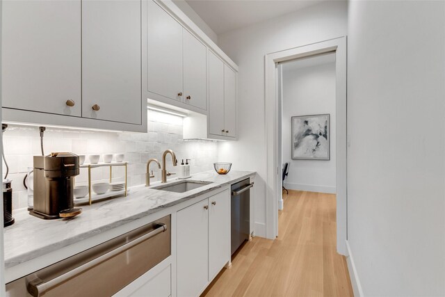 kitchen featuring light wood finished floors, stainless steel dishwasher, a sink, tasteful backsplash, and light stone countertops