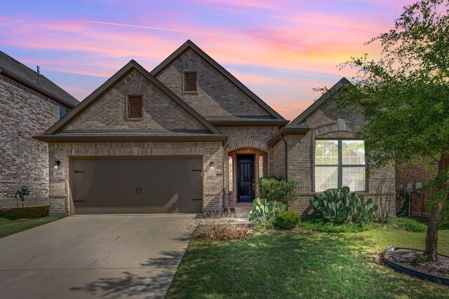 french country inspired facade featuring concrete driveway, a lawn, an attached garage, and brick siding
