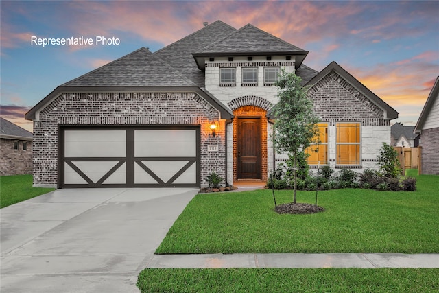 french country home featuring a shingled roof, concrete driveway, a lawn, brick siding, and a garage