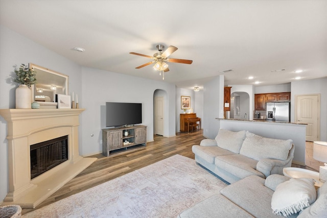living room with a fireplace with raised hearth, light wood-style floors, ceiling fan, arched walkways, and visible vents