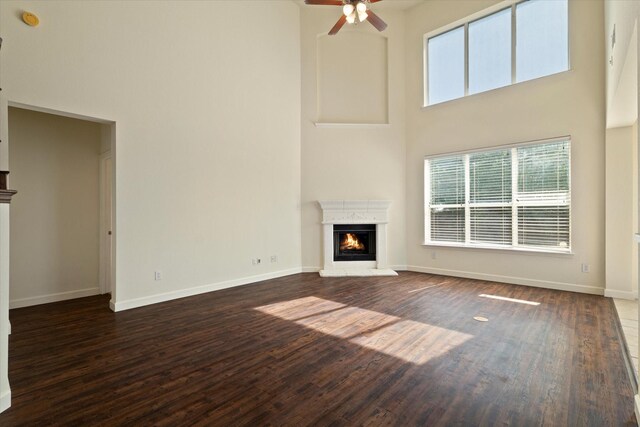 unfurnished living room with dark wood finished floors, a warm lit fireplace, a towering ceiling, and a ceiling fan