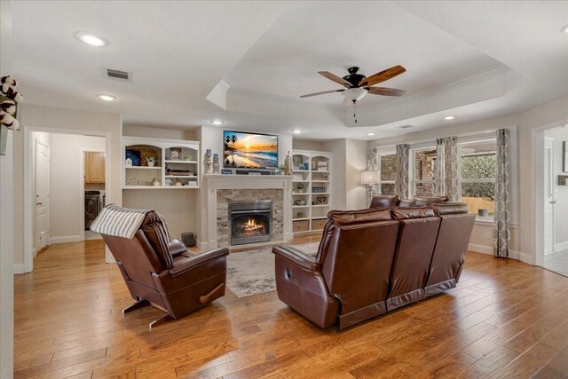 living area with a warm lit fireplace, ceiling fan, light wood finished floors, a raised ceiling, and visible vents