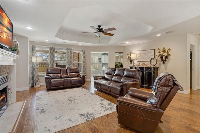 living room featuring wood finished floors, a warm lit fireplace, ceiling fan, a tray ceiling, and baseboards
