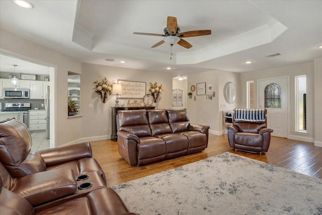 living room featuring ceiling fan, a tray ceiling, light wood-style flooring, ornamental molding, and visible vents