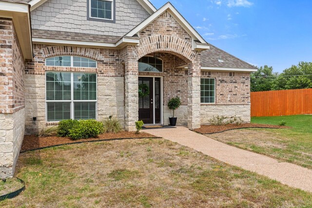 doorway to property featuring fence, a lawn, brick siding, and roof with shingles