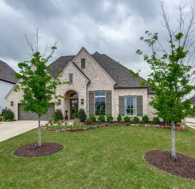french country home with a shingled roof, driveway, a front lawn, and brick siding