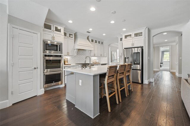kitchen featuring a kitchen island with sink, stainless steel appliances, arched walkways, and custom range hood