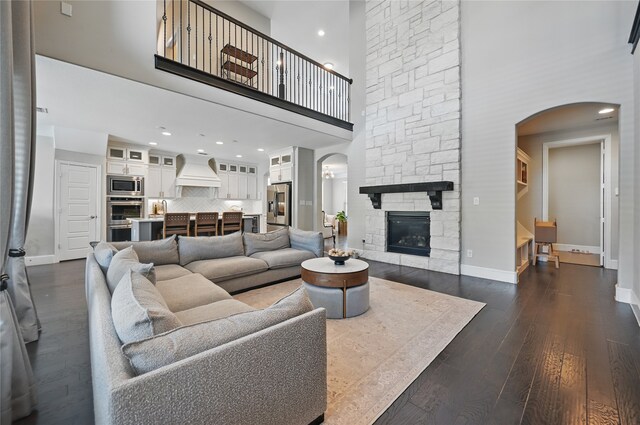 living room featuring arched walkways, dark wood-style floors, a stone fireplace, and a towering ceiling