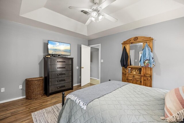 bedroom featuring a tray ceiling, visible vents, baseboards, a ceiling fan, and wood finished floors