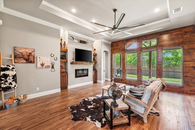 living area with a raised ceiling, a glass covered fireplace, visible vents, a ceiling fan, and ornamental molding