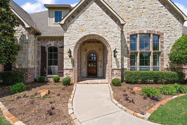 view of exterior entry with stone siding, a shingled roof, and brick siding