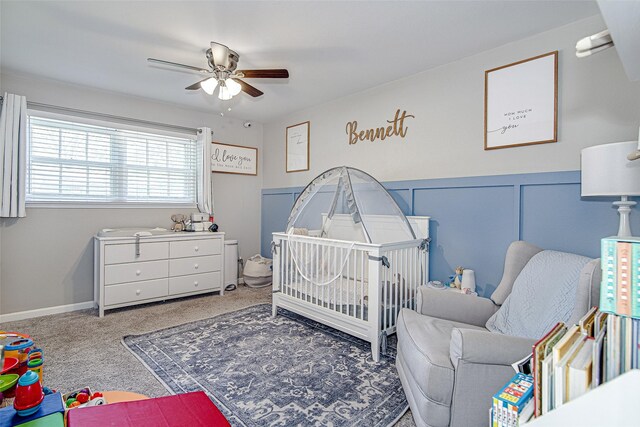 bedroom featuring wainscoting, a nursery area, carpet floors, and ceiling fan