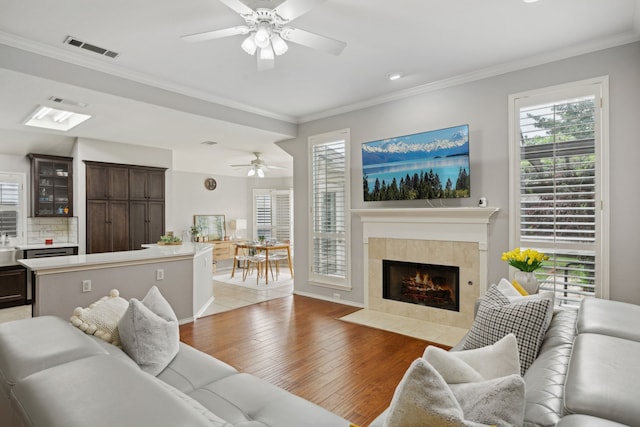 living area featuring crown molding, visible vents, ceiling fan, light wood-type flooring, and a tiled fireplace
