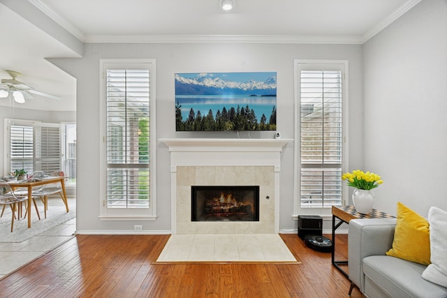 living area featuring a tiled fireplace, wood-type flooring, crown molding, a ceiling fan, and baseboards