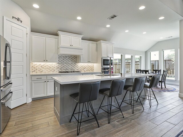 kitchen with stainless steel appliances, a kitchen island with sink, light wood-type flooring, visible vents, and lofted ceiling