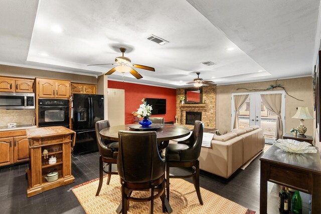 dining space featuring a tray ceiling, dark wood-style floors, visible vents, a brick fireplace, and ceiling fan