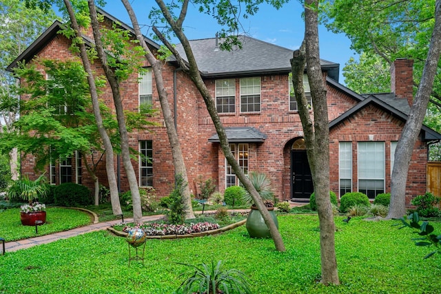 tudor home featuring brick siding, roof with shingles, a chimney, and a front lawn