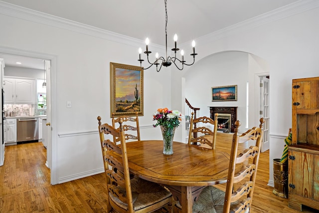 dining space with light wood finished floors, ornamental molding, arched walkways, a lit fireplace, and a notable chandelier