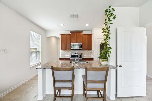 kitchen with brown cabinets, stainless steel appliances, light stone countertops, decorative backsplash, and a breakfast bar area
