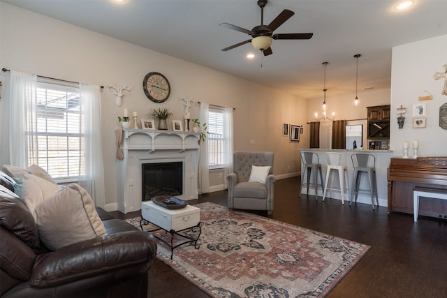 living area with a glass covered fireplace, baseboards, dark wood-style flooring, and ceiling fan with notable chandelier