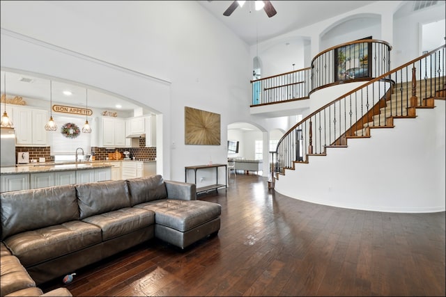 living room with arched walkways, dark wood-style flooring, a towering ceiling, stairs, and ceiling fan