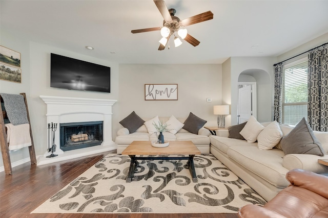 living area with a fireplace with raised hearth, baseboards, arched walkways, wood finished floors, and a ceiling fan