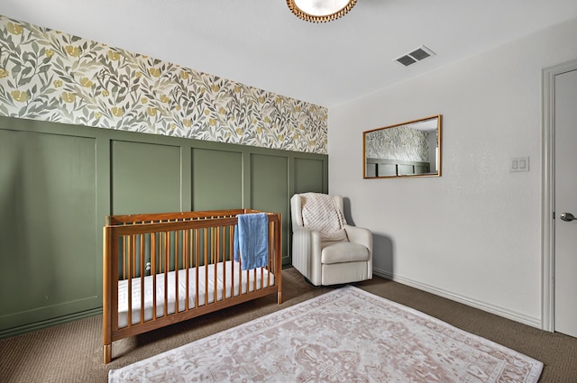 carpeted bedroom featuring visible vents, a wainscoted wall, a nursery area, wallpapered walls, and baseboards