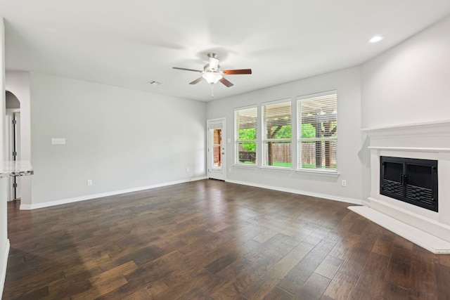 unfurnished living room featuring dark wood-style floors, a fireplace with raised hearth, a ceiling fan, baseboards, and arched walkways