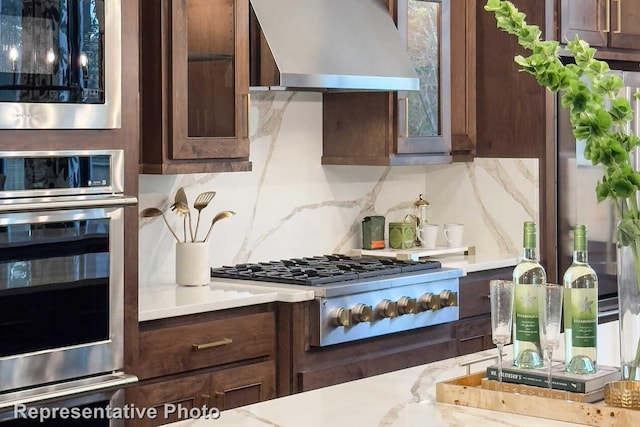 kitchen featuring appliances with stainless steel finishes, glass insert cabinets, wall chimney exhaust hood, backsplash, and dark brown cabinets