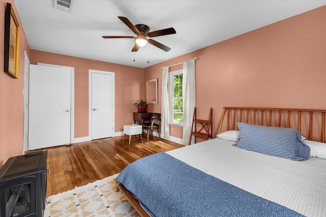 bedroom with a wood stove, a ceiling fan, visible vents, baseboards, and wood finished floors