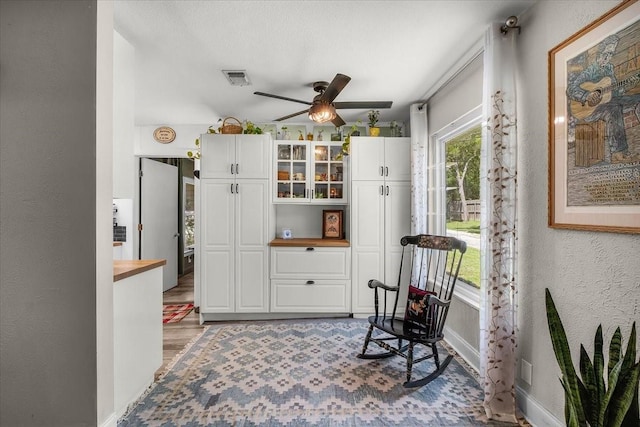 living area featuring light wood finished floors, a textured wall, baseboards, visible vents, and a ceiling fan