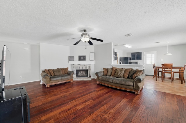 living room with a textured ceiling, ceiling fan, visible vents, wood finished floors, and a stone fireplace