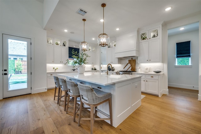 kitchen featuring white cabinetry, decorative backsplash, custom range hood, visible vents, and a kitchen island with sink
