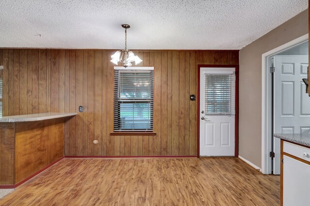 unfurnished dining area featuring a textured ceiling, light wood finished floors, baseboards, a notable chandelier, and wooden walls