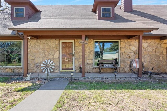 doorway to property with a chimney, a yard, covered porch, stone siding, and roof with shingles
