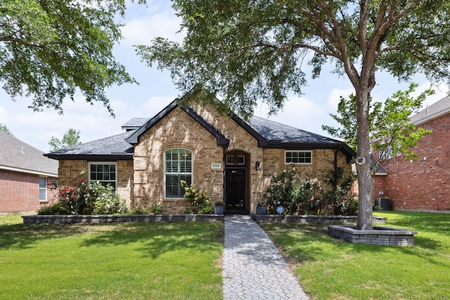 tudor-style house with a shingled roof, cooling unit, a front lawn, and brick siding